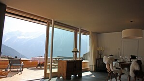 Een kamer met een groot raam met uitzicht op bergen, een houten eettafel en een gezellige zithoek met stoelen.