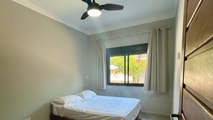 Un dormitorio con una cama individual, ventilador de techo, ventana con cortinas y vista a espacios verdes.