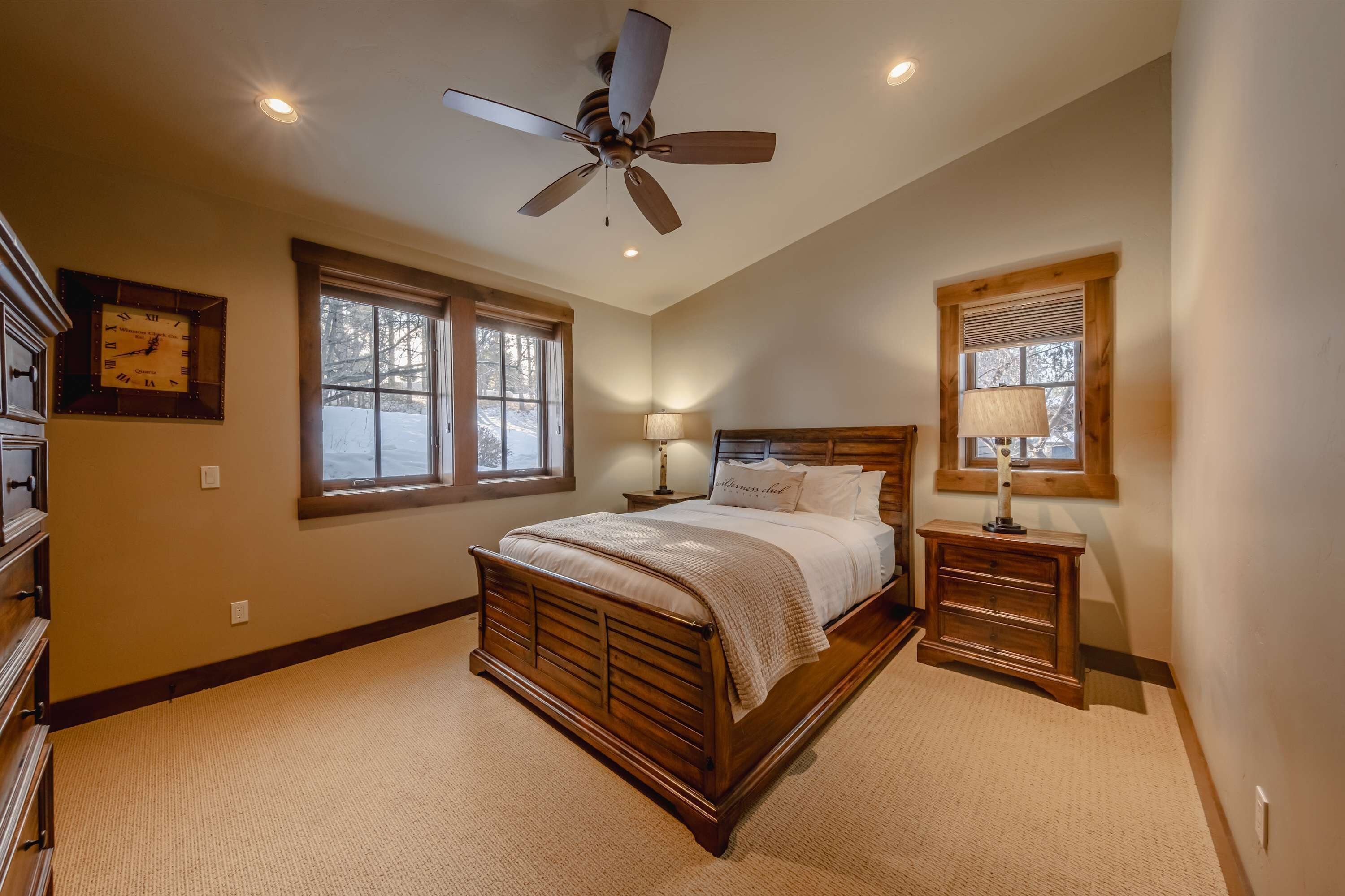 A bedroom with a wooden bed, two bedside tables, a ceiling fan, and two windows.