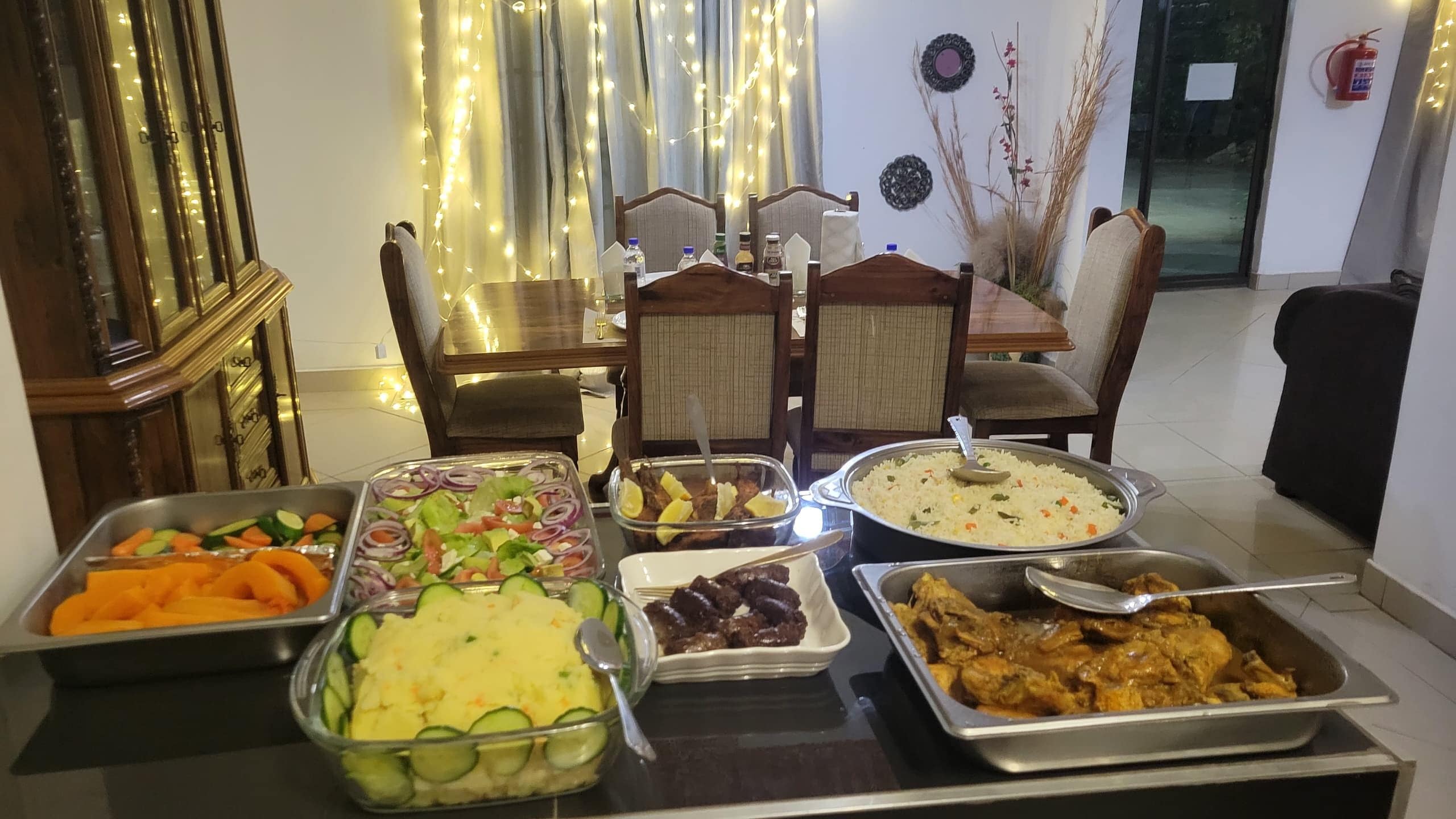 Una mesa de buffet con diversos platos, una zona de comedor con sillas y una pared decorativa con luces.