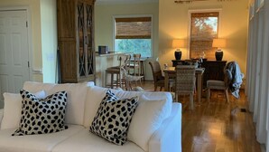 Un salón con un sofá blanco, dos almohadas estampadas, una mesa de centro de madera y un comedor con sillas y una mesa.