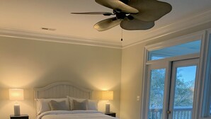 Un dormitorio con cama, dos mesitas de noche con lámparas, ventilador de techo y vistas a través de un ventanal corredero.