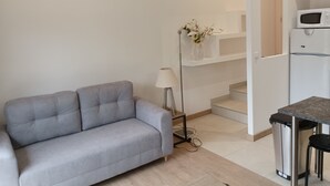 Een moderne woonkamer met een grijze bank, een vloerlamp en een klein tafeltje. Een trap leidt naar een keuken met een magnetron en een prullenbak.