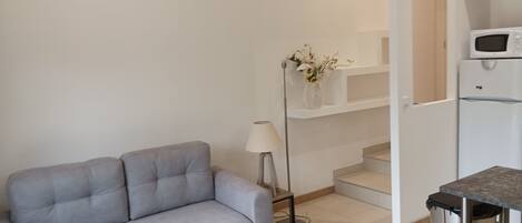 A modern living room with a grey sofa, a floor lamp, and a small table. A staircase leads to a kitchen area with a microwave and a trash bin.