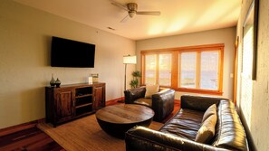 A living room with a flat-screen TV mounted on the wall, a wooden entertainment unit, leather sofas, a round coffee table, and a ceiling fan. - 2-Bedroom, 1.5-Bathroom Suite at REAL Watersports (Waves)