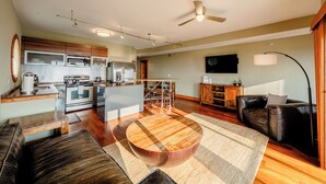 A modern kitchen with stainless steel appliances, a dining area with a wooden table and stools, a leather sofa, and a flat-screen TV mounted on the wall. - Waterfront 2-Bedroom, 1.5-Bathroom Suite at REAL Watersports (Waves)