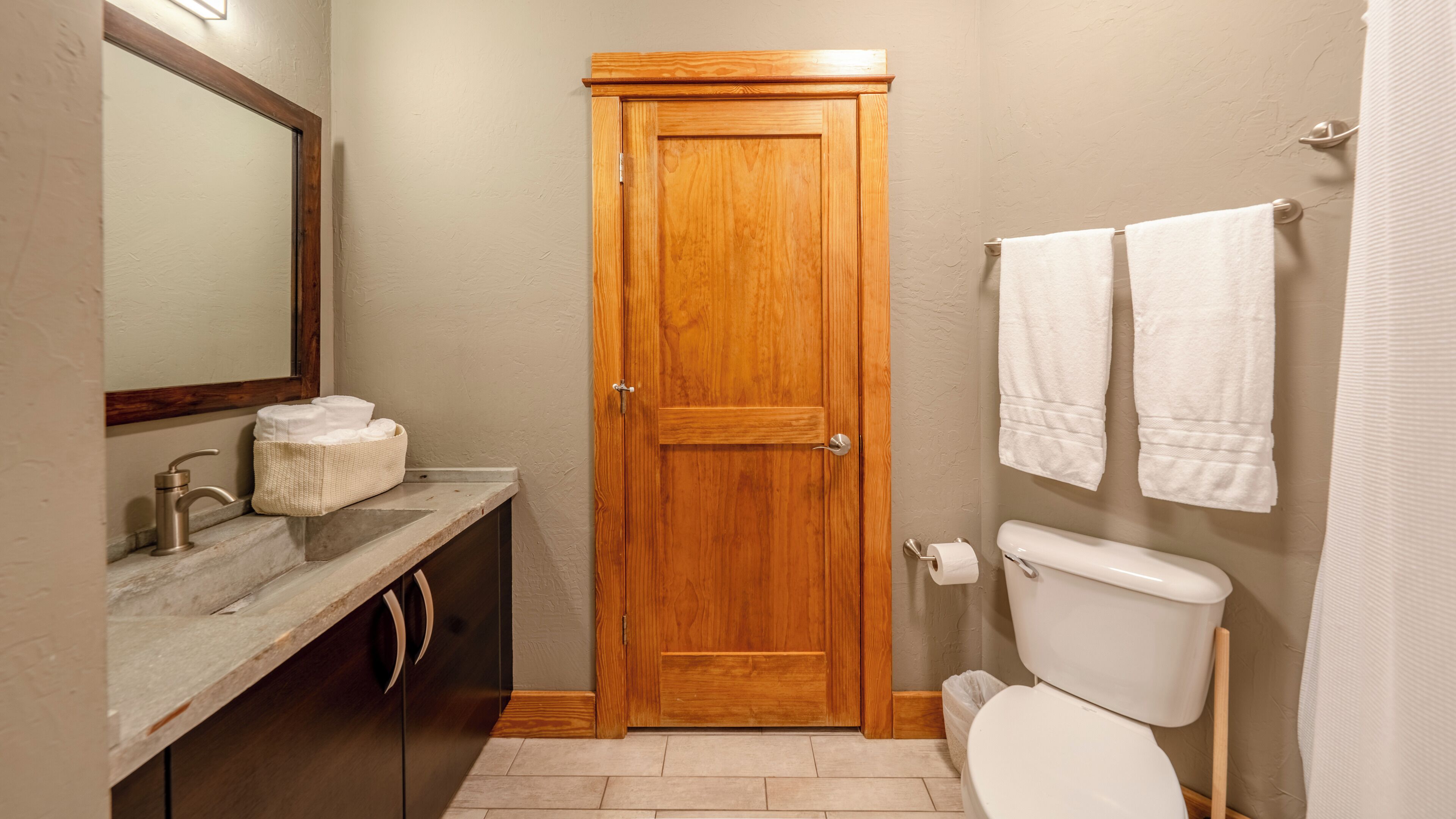 A bathroom with a wooden door, a toilet, a sink with a towel dispenser, and two towels hanging on the wall.