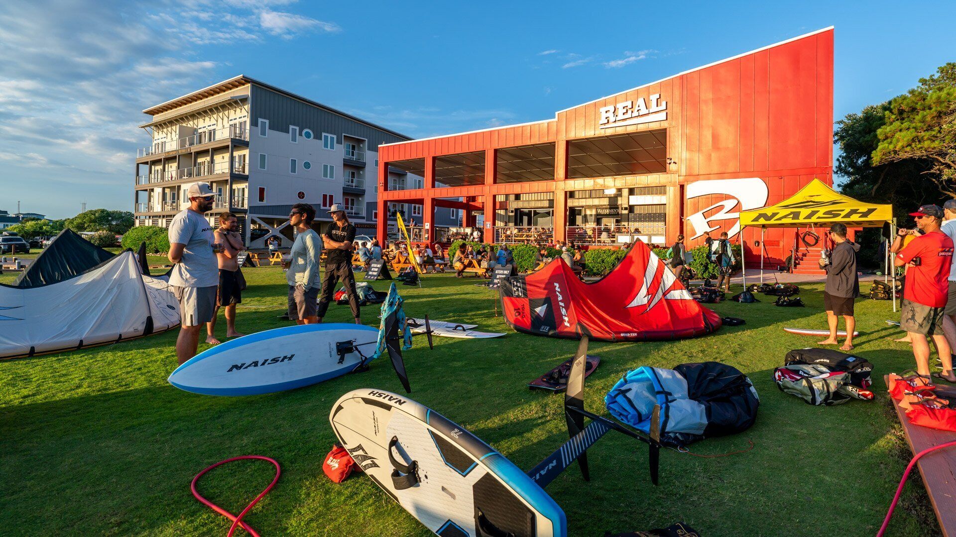 A building with a 'REAL' sign, a tent, and several surfboards on the grass.