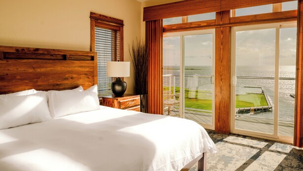 A hotel room with a large bed, a nightstand with a lamp, and a view of the sea through a sliding glass door. - Waterfront 3-Bedroom, 2.5-Bathroom Penthouse Suite at REAL Watersports (Waves)