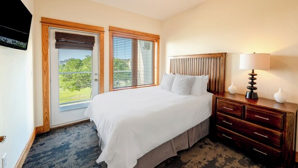 A hotel room with a large bed, wooden headboard, a nightstand with a lamp, and a view of greenery through the windows. - Waterfront 3-Bedroom, 2.5-Bathroom Penthouse Suite at REAL Watersports (Waves)