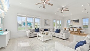 Amplia sala de estar con sofá seccional, mesa de centro y comedor con mesa y sillas de madera. Hay ventiladores de techo, ventanas grandes y vistas al exterior.
