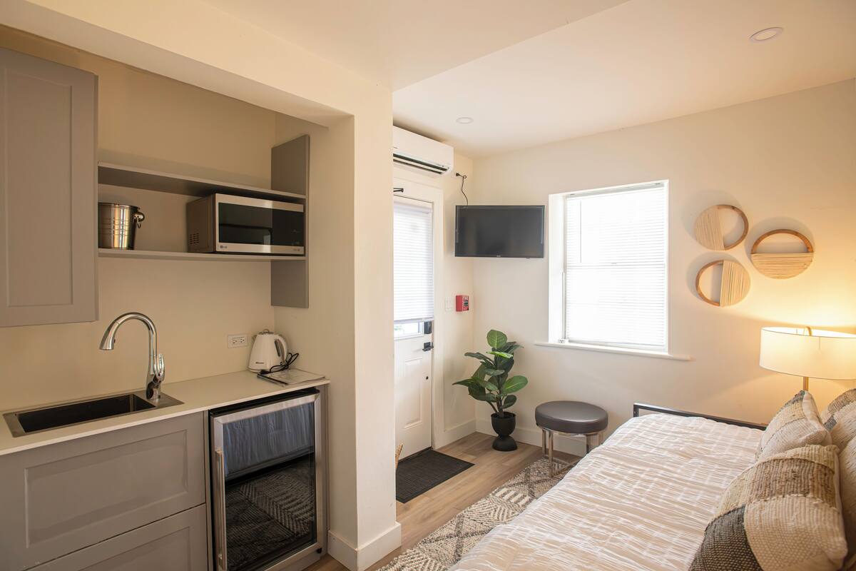 Una cocina compacta con microondas, pileta y horno, un pequeño comedor con una mesa y una silla, una planta en maceta y una cama con mesita de noche y lámpara.