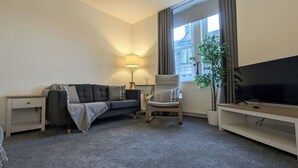 Ein modernes Wohnzimmer mit einer grauen Couch, einem Holzstuhl, einem Fernseher und einem großen Fenster mit Vorhängen.