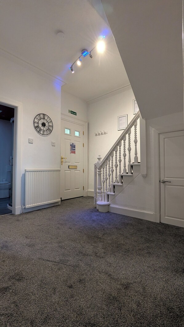 A hallway with a white door, a fire exit sign, a clock, and a staircase with white balusters.