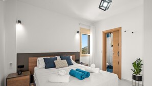 Ein modernes Hotelzimmer mit einem großen Bett, Nachttischen, einer Nachttischlampe, einer Topfpflanze und Blick ins Freie.
