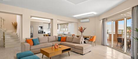 A modern living room with a sectional sofa, a coffee table, and a dining area with chairs and a table. There is a staircase leading to an upper level, and a kitchen area is visible in the background.