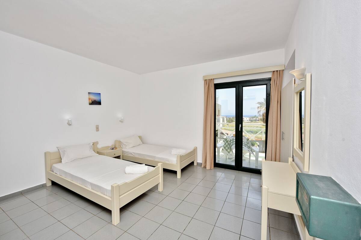 Une chambre avec deux lits simples, un bureau, une chaise, une fenêtre avec des rideaux et une vue sur l’extérieur.