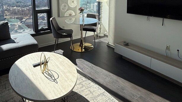 A modern living room with a large window, a round coffee table, a sofa, a TV mounted on the wall, and a dining area with chairs and a table. - Stylish King Suite • Tysons Corner Mall Access • Metro Nearby (Tysons)