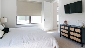 Une chambre à coucher comprenant un lit, une commode, un téléviseur et une fenêtre avec des stores.