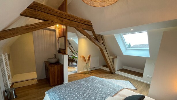 A bedroom with a sloped ceiling, a bed with patterned bedding, a wooden dresser, a small table with a bowl, and a view of a staircase and a window. - La Girouette - in the heart of a farmhouse (Mittelhausbergen)