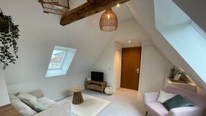 A modern loft apartment with a high ceiling, exposed wooden beams, a cozy seating area with a sofa and armchair, a small round table, and a television on a wooden stand. - La Girouette - in the heart of a farmhouse (Mittelhausbergen)