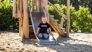 Een houten speelconstructie met een glijbaan en klimtrappen, omgeven door zand en begroeiing.