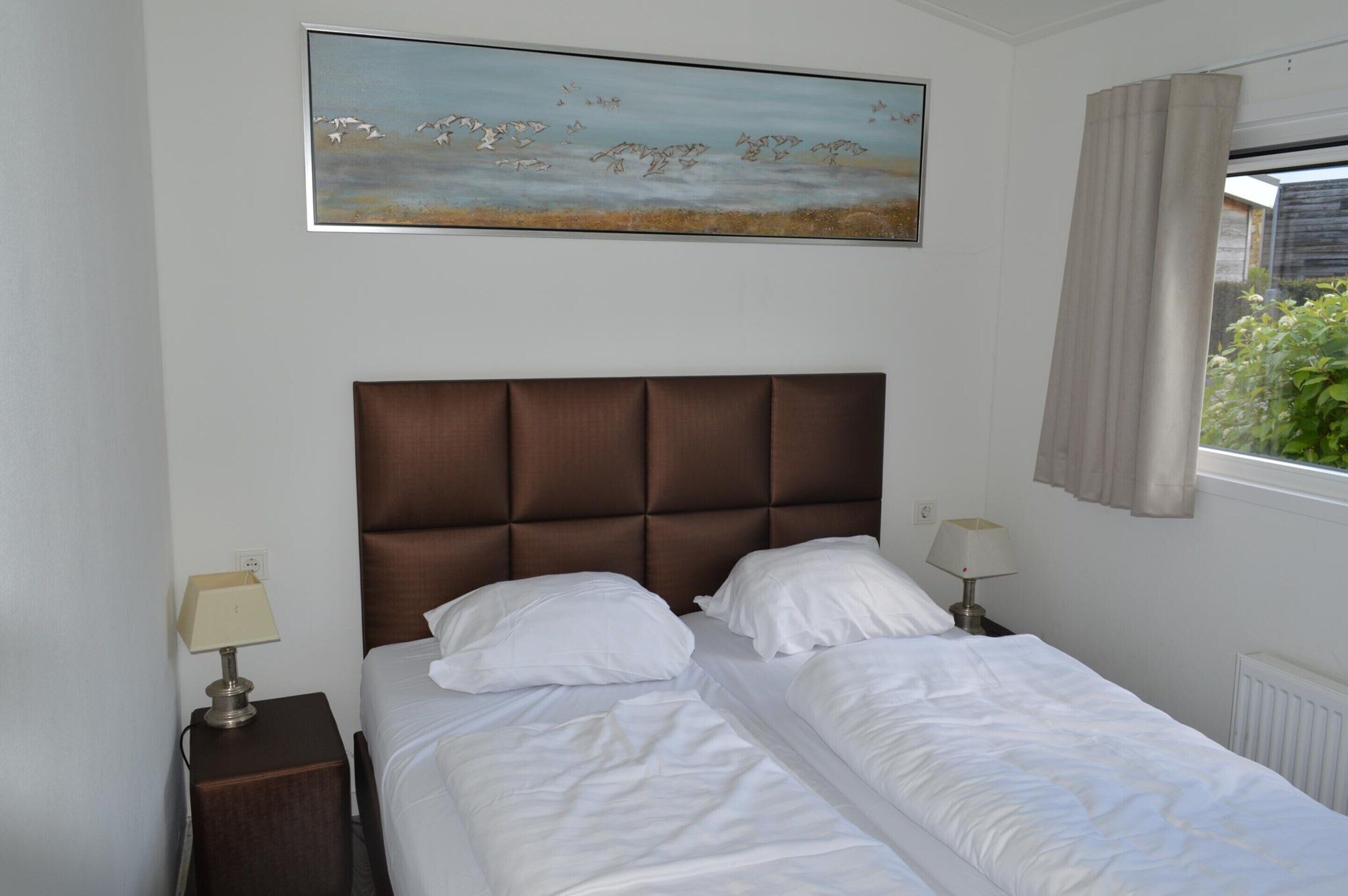Een keurig opgemaakt bed met witte lakens, een bruin bekleed hoofdbord en een schilderij van vogels aan de muur.