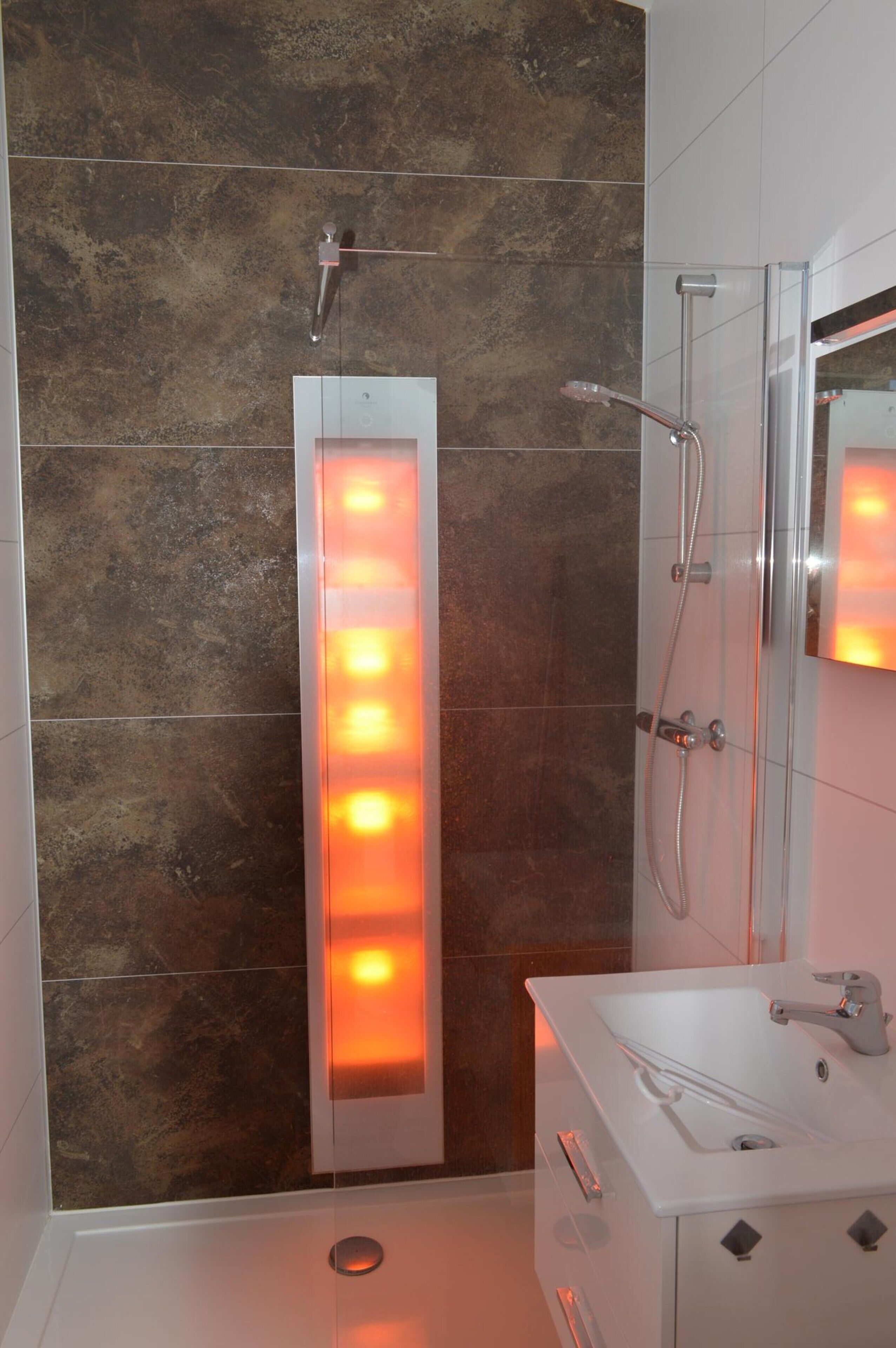Une salle de bain moderne avec un panneau lumineux vertical, une cabine de douche en verre et un lavabo blanc avec un miroir.