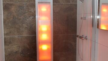 Une salle de bain moderne avec un panneau lumineux vertical, une cabine de douche en verre et un lavabo blanc avec un miroir.