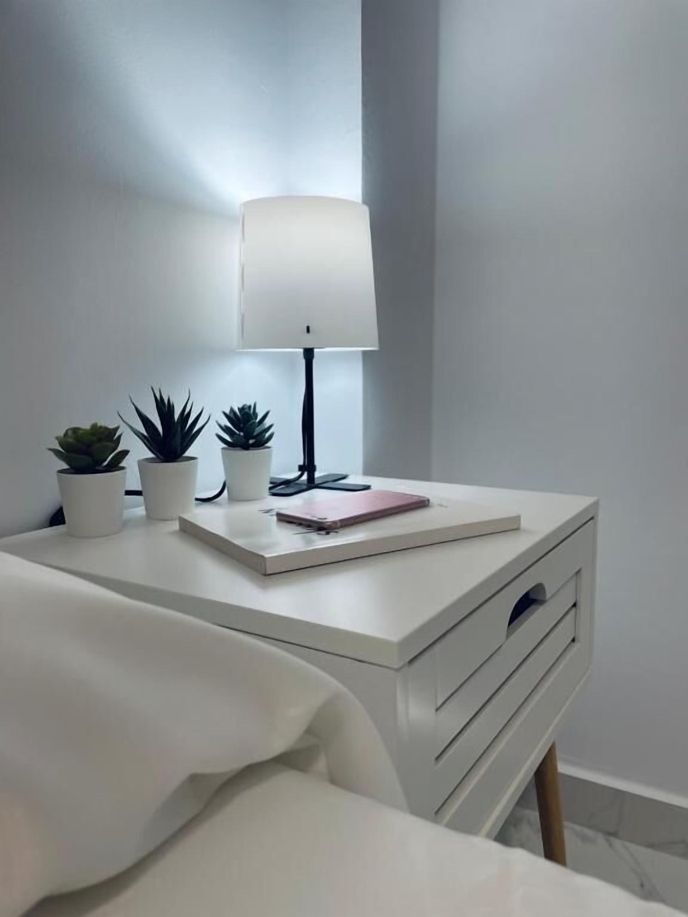 Et minimalistisk natbord med en lampe, to krukker med planter og en notesbog.
