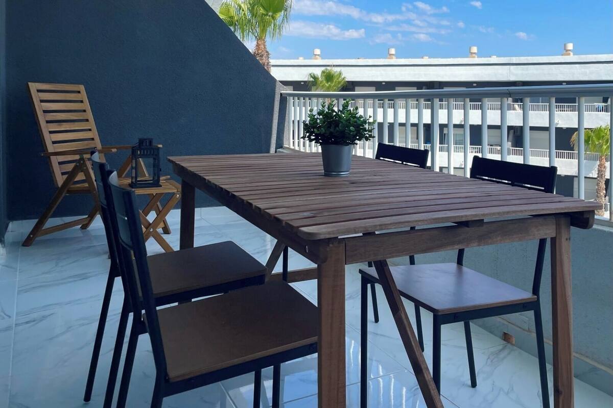 Una mesa de madera con cuatro sillas en un balcón que ofrece vistas a un edificio y palmeras.