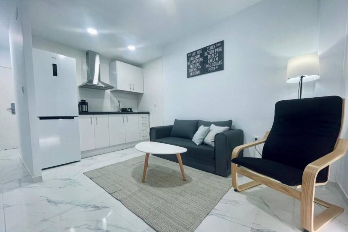 Una sala de estar moderna con un sofá, una mesa de centro y una silla. Hay una zona de cocina con armarios y un refrigerador. Se ve un letrero con texto en la pared.