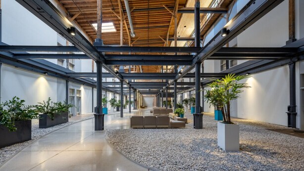 Un espacio interior amplio con techo alto, vigas metálicas y una zona de estar central con sofás.