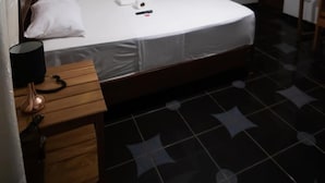 A single bed with a wooden headboard, a bedside table with a lamp, a remote control, and a white pillow. The room has a tiled floor with a star pattern, beige curtains, and a hanging light fixture. - Puerto Azul  (Sauce)