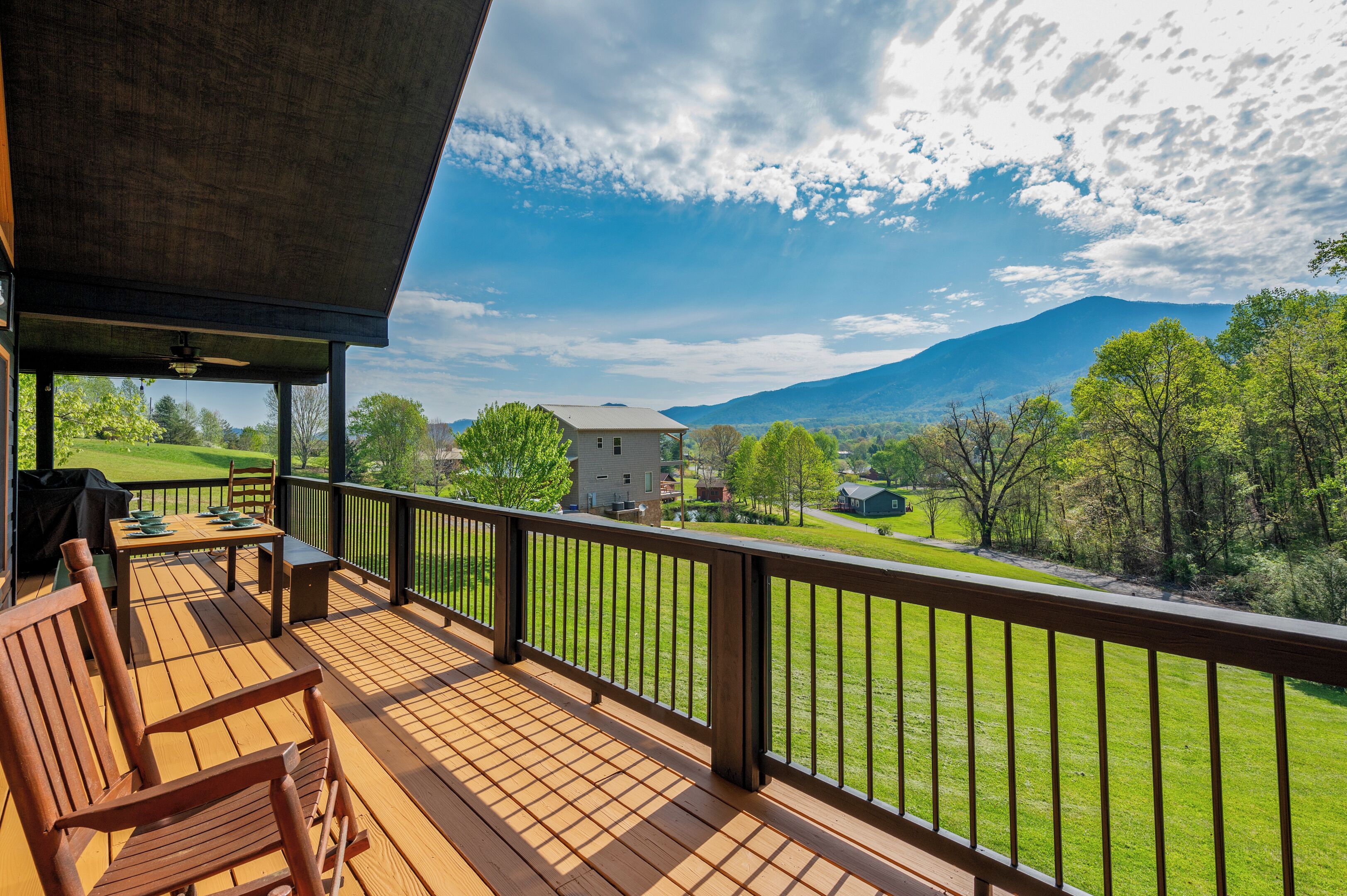 A wooden deck with a table and chairs, overlooking a green landscape with trees and a mountain in the distance.