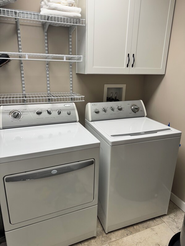 A laundry room with two stacked washer and dryer units, a metal shelf above, and a white cabinet below.