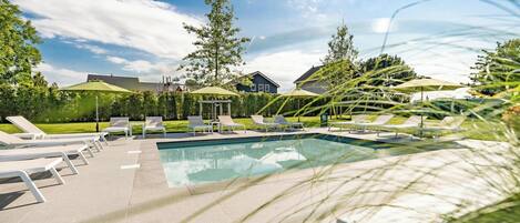 A swimming pool with lounge chairs and umbrellas, surrounded by greenery and houses.