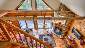 A wooden interior with a stone fireplace, a wooden staircase, and a view of the outdoors through large windows. - Slopeside | Hocking Hills Retreat, Covered Hot Tub (Laurelville)