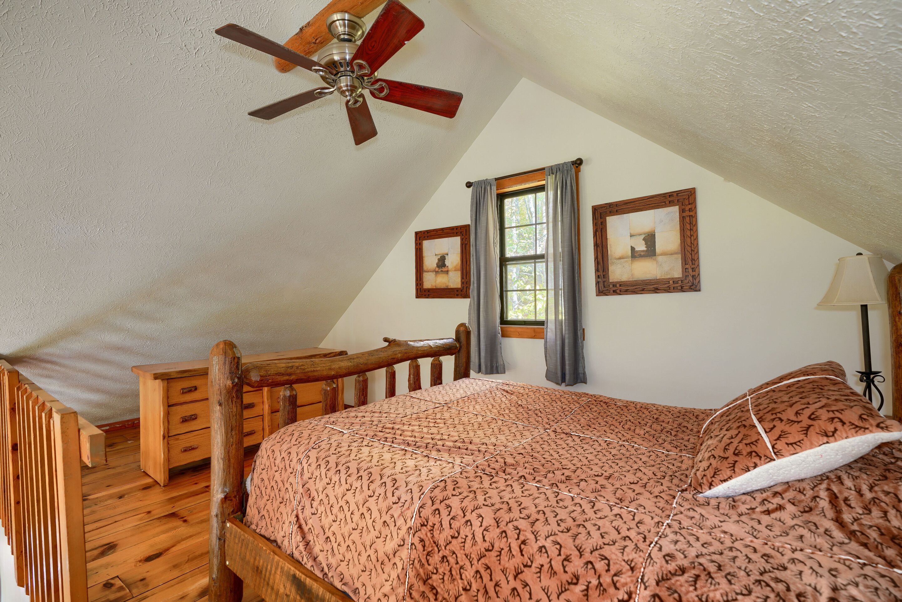 A bedroom with a sloped ceiling, a wooden bed frame, a ceiling fan, two framed pictures, a window with curtains, and a bedside lamp.