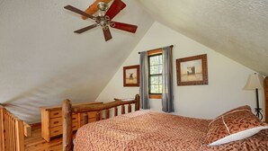 A bedroom with a sloped ceiling, a wooden bed frame, a ceiling fan, two framed pictures, a window with curtains, and a bedside lamp. - Slopeside | Hocking Hills Retreat, Covered Hot Tub (Laurelville)