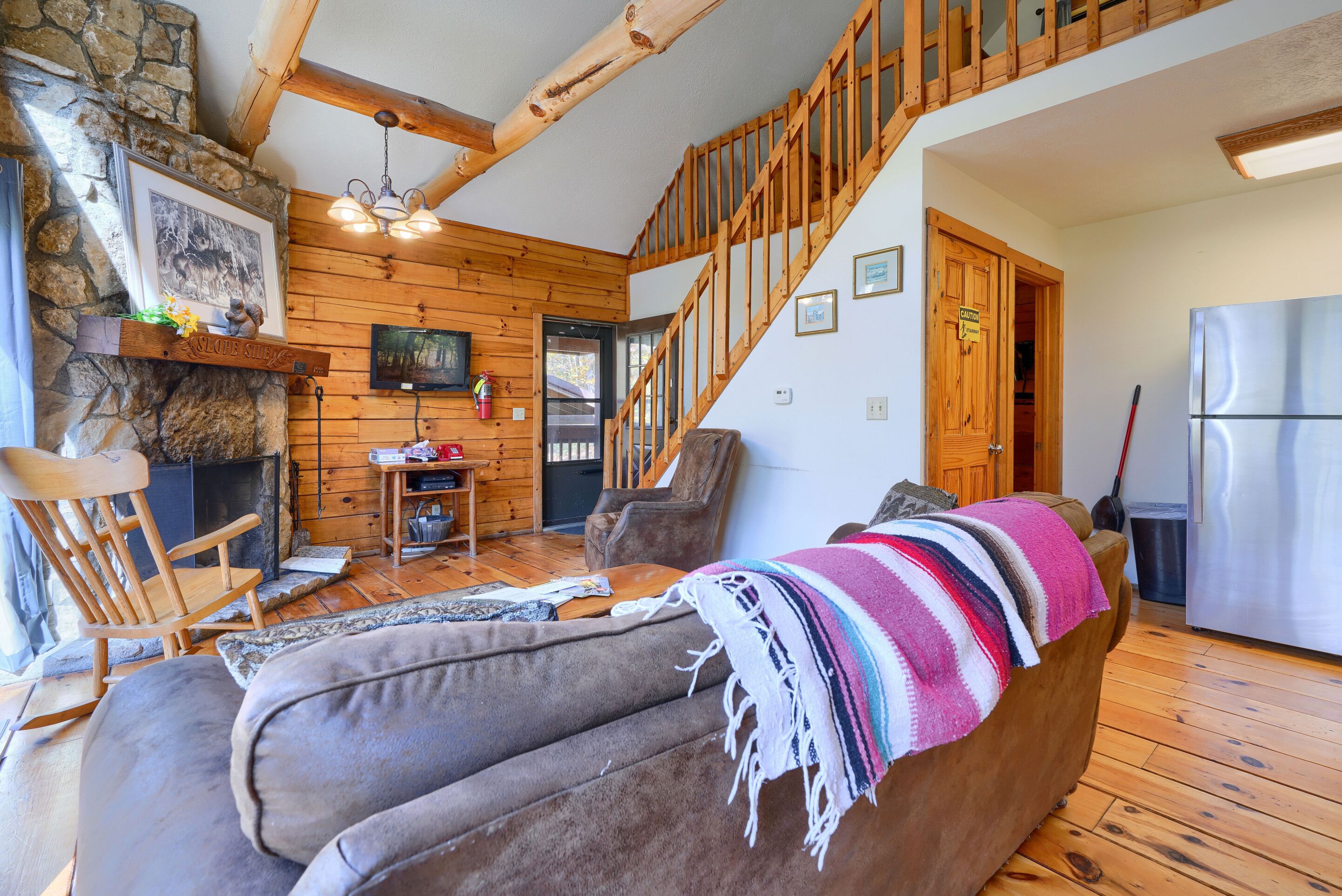 A cozy living room with a stone fireplace, wooden ceiling beams, a staircase, and a modern kitchen.