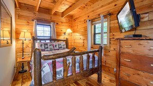 Ein Schlafzimmer einer Holzhütte mit Bett, Schminktisch, Fernseher und zwei Fenstern.