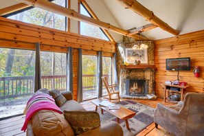 A cozy cabin interior with a stone fireplace, wooden walls, and large windows offering a view of the surrounding forest.