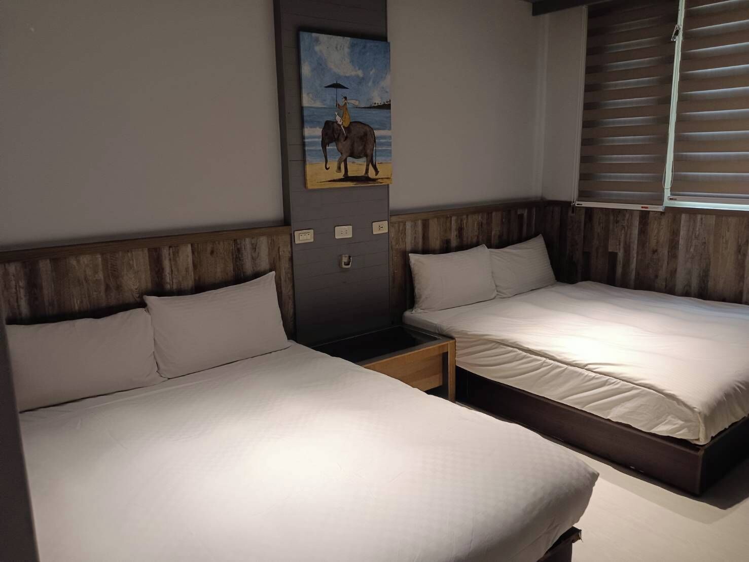 A hotel room with two beds, wooden headboards, a nightstand, a painting of an elephant, and a window with blinds.