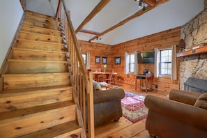 Eine Holztreppe führt zu einem Wohnbereich mit Kamin aus Stein, Holzwänden und einem Esstisch.
