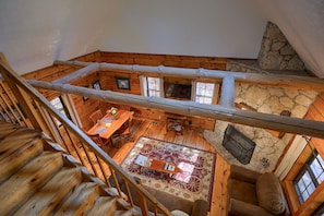 A wooden cabin interior with a loft bedroom, a stone fireplace, and a dining area.