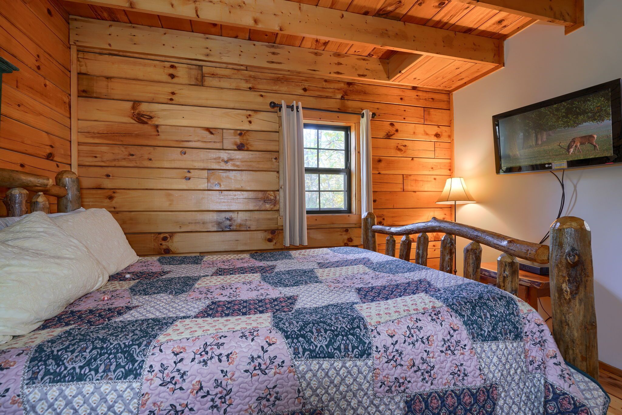 Una habitación de cabaña de madera con una cama, una ventana con cortinas, una lámpara y un cuadro en la pared.