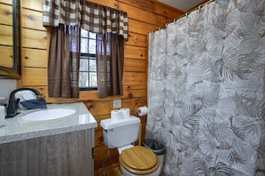 Une salle de bain rustique, avec des murs en bois, comprenant des toilettes en bois, un lavabo et une fenêtre avec des rideaux.