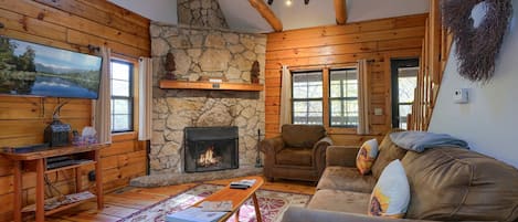 A cozy living room with wooden walls, a stone fireplace, a leather sofa, and a large window.
