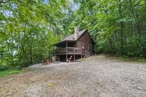 Une cabane en rondins dotée d’un porche et d’une terrasse, entourée d’une forêt dense.